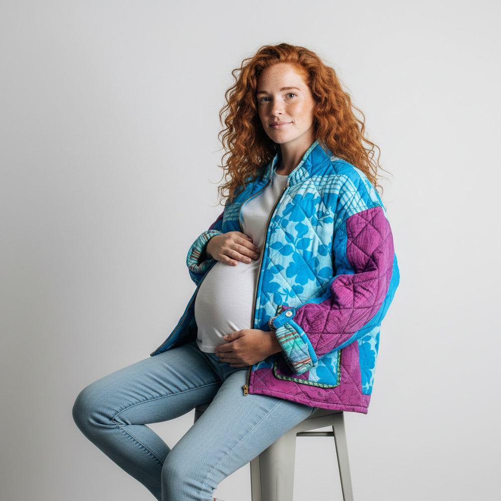 Pregnant model wearing a colorful quilted jacket sitting on a stool against a plain background